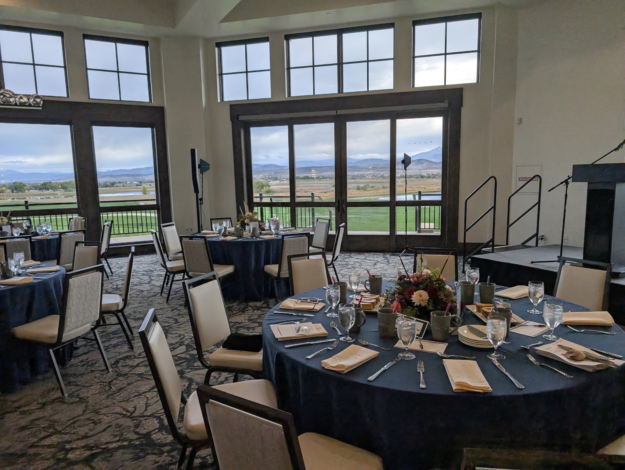 A banquet room set for an event, with speaker columns on stands by the windows and a stage at right